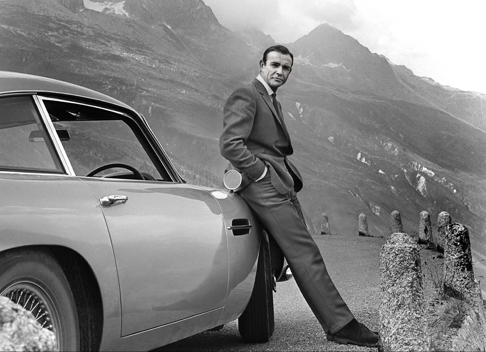 Sean Connery, starring as James Bond in Goldfinger, leans against his Aston Martin DB5 silver birch colored car