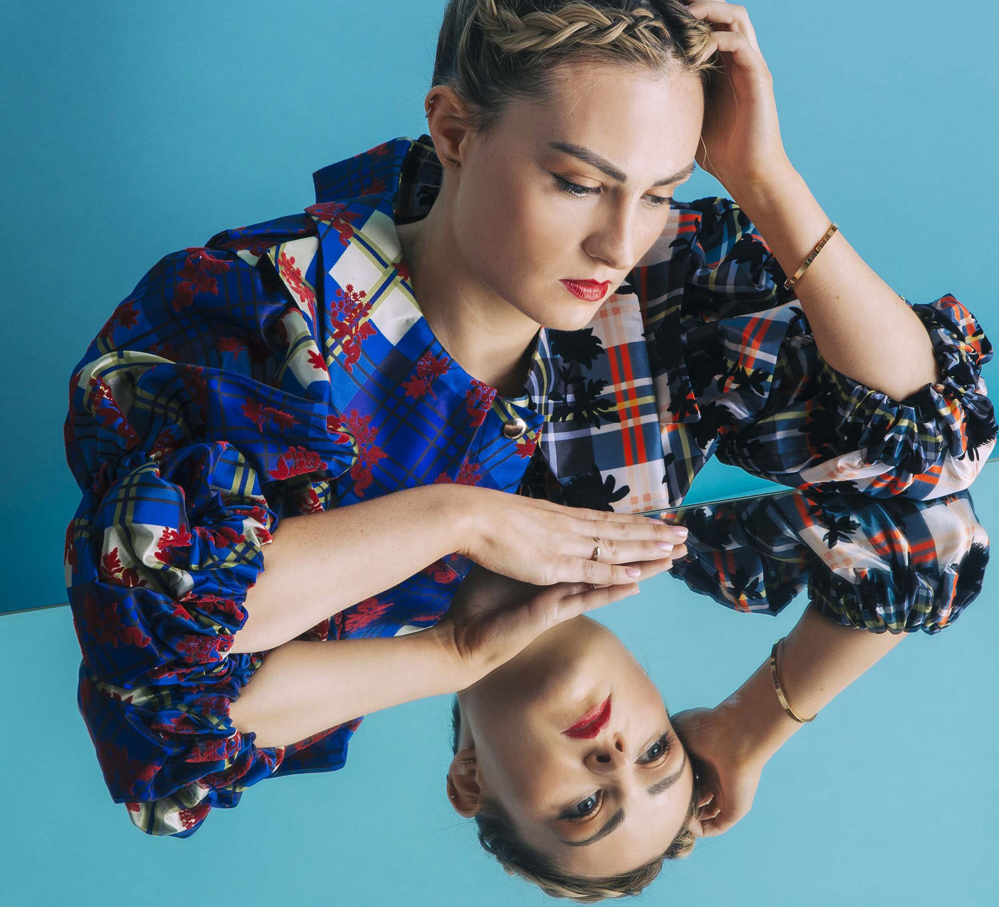 Molly is sitting down at a table with a mirrored surface. You can see her reflection in the mirror. Her hair is in an up-do with braids and a bun. She is wearing a printed multi-colour blouse.